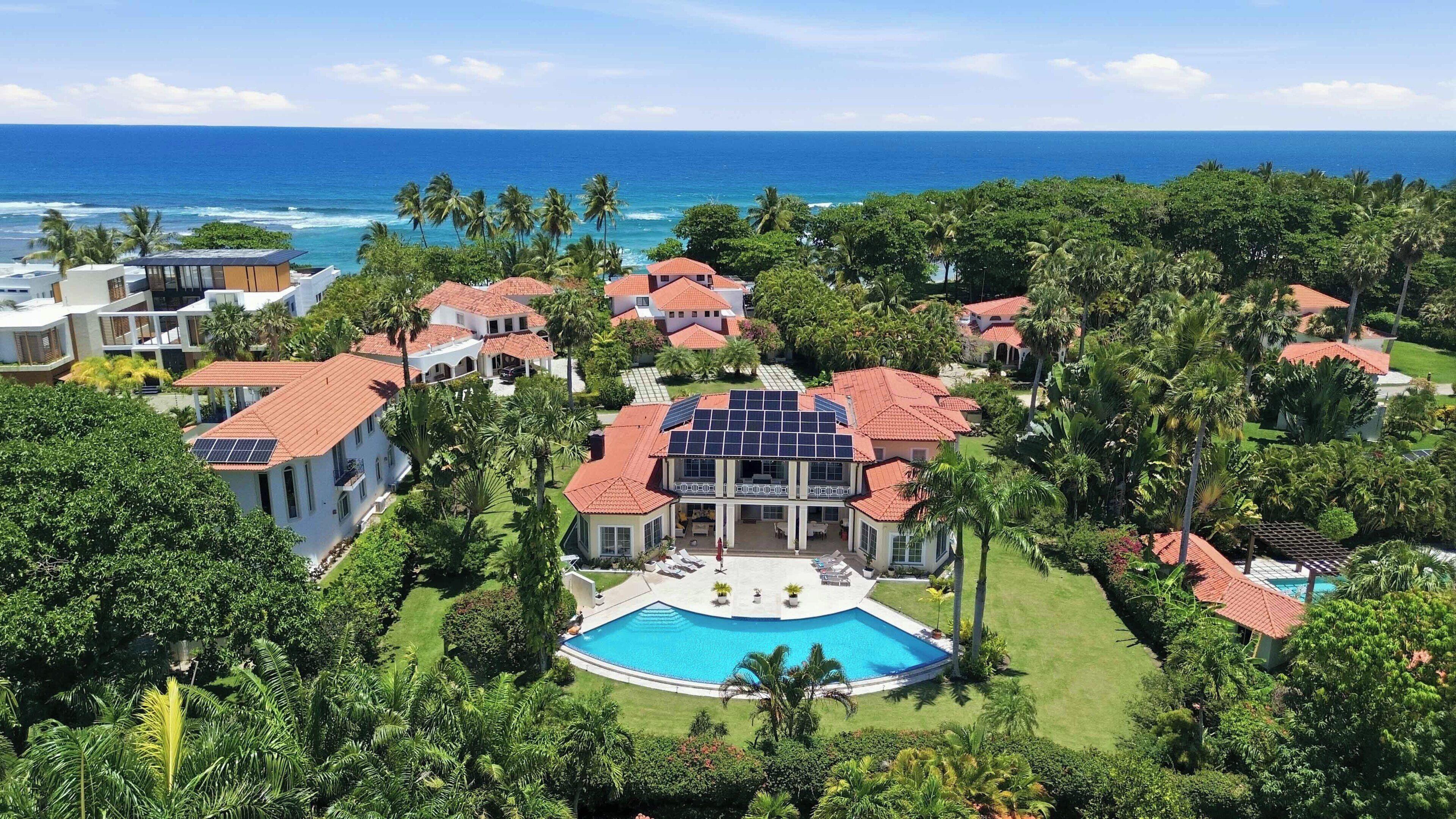 A resort with a large swimming pool, multiple buildings with red-tiled roofs, and a view of the ocean.