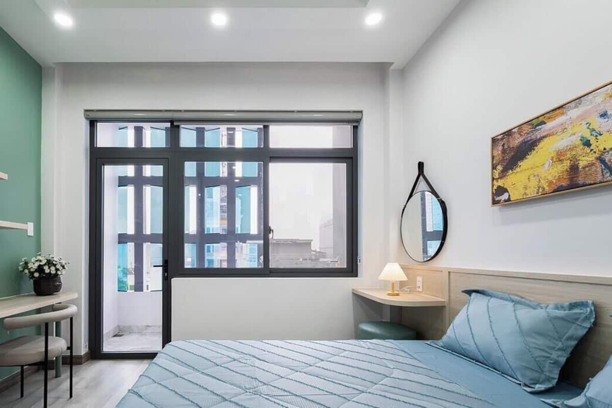 A bedroom with a bed, a desk, a chair, a window, a bedside table, a lamp, and a painting on the wall.