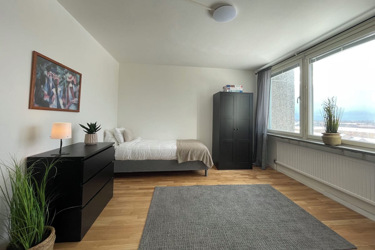 Un dormitorio con cama, cómoda, aparador, lámpara, planta y ventana con vista.