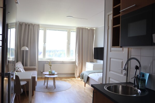 A modern hotel room with a bed, a desk, a dining area, a kitchen with a sink, and a large window.