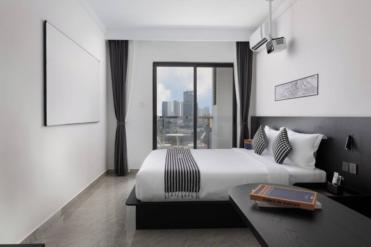 A modern hotel room with a large bed, a city view, a projector, and a map on the wall.