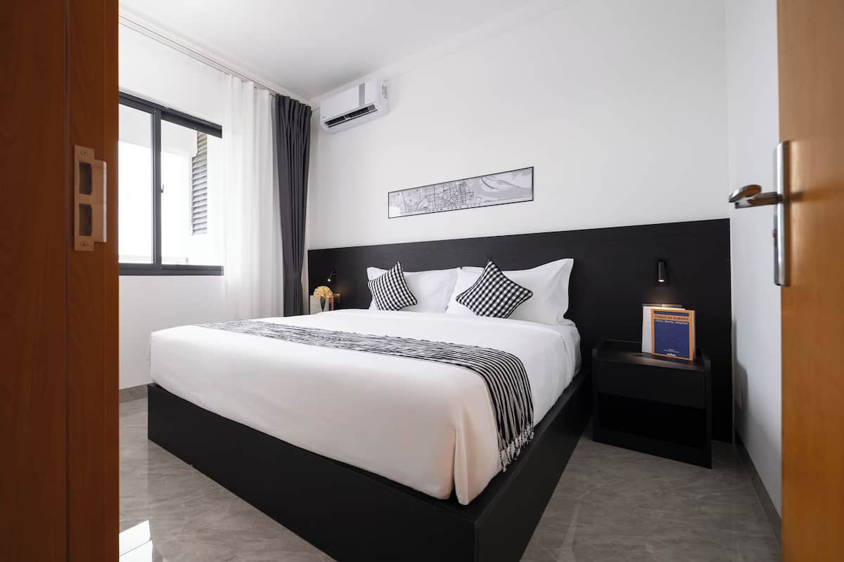 A modern hotel room with a large bed, a nightstand, a wall-mounted air conditioner, and a window with curtains.