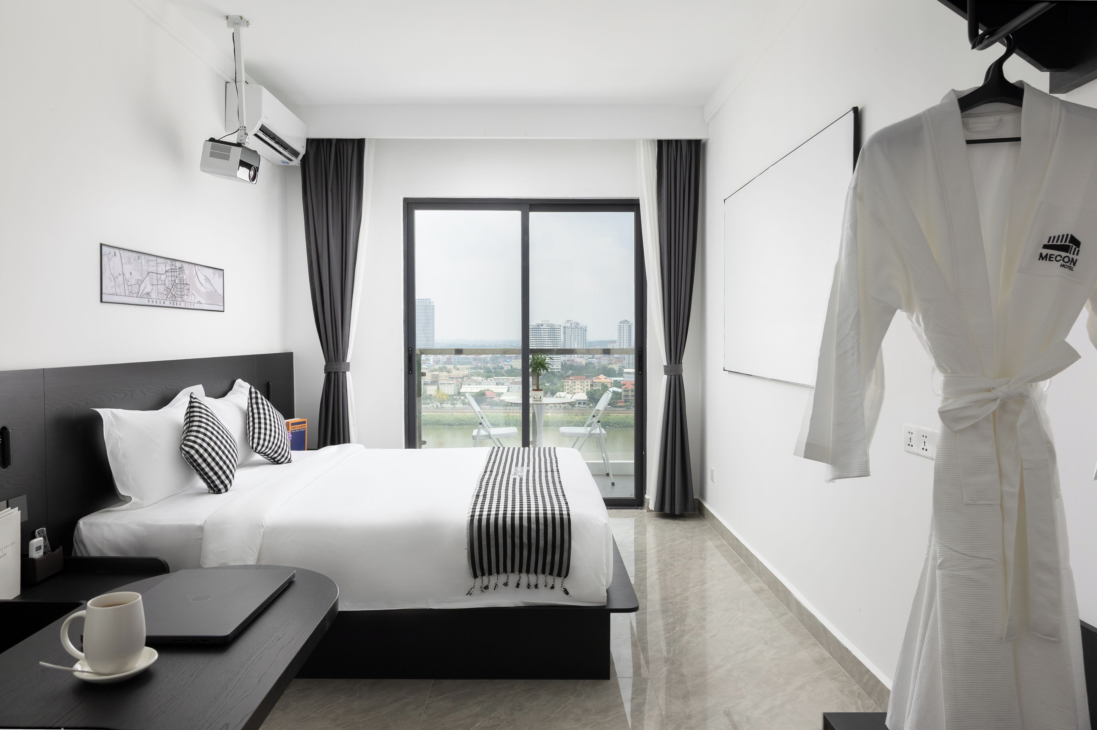 A hotel room with a bed, a desk with a laptop and a cup, a robe hanging on the wall, and a large window with a city view.