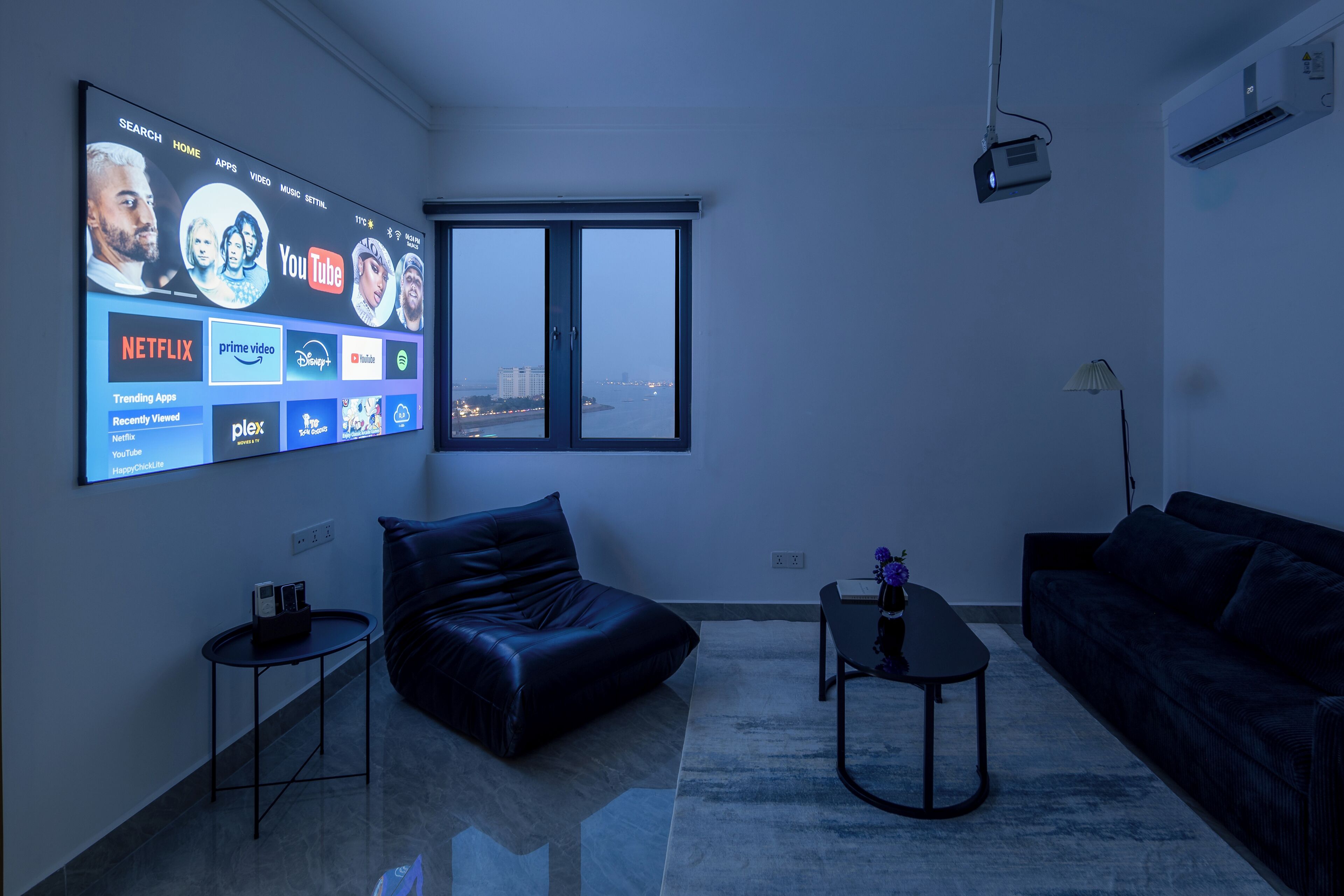 Een moderne woonkamer met een grote tv waarop streamingdiensten worden afgespeeld, een leren bank, een salontafel en een uitzicht op een stadsgezicht door een raam.