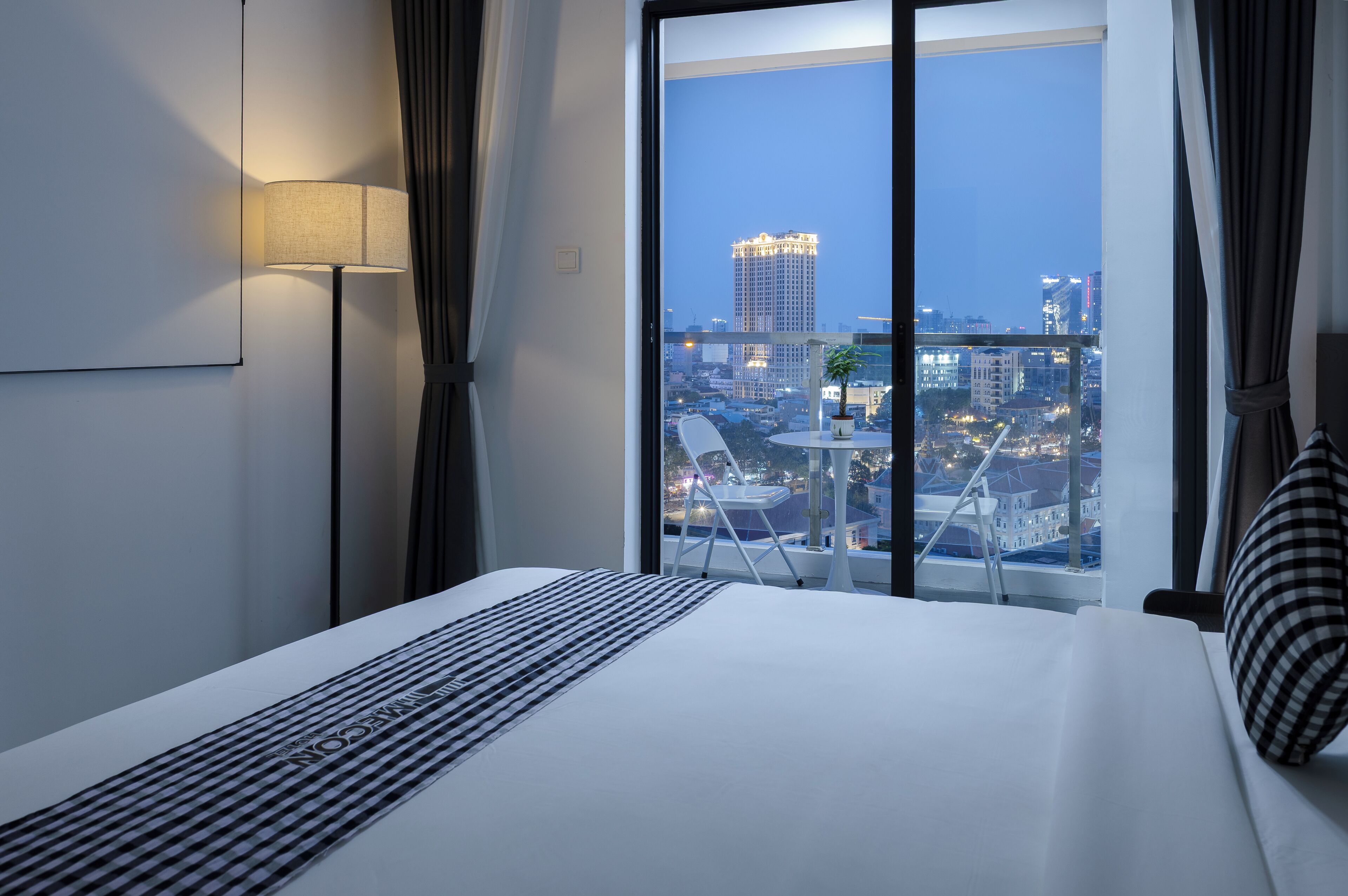 Habitación de hotel con cama, lámpara, vista del paisaje urbano a través de un ventanal grande y una silla fuera.