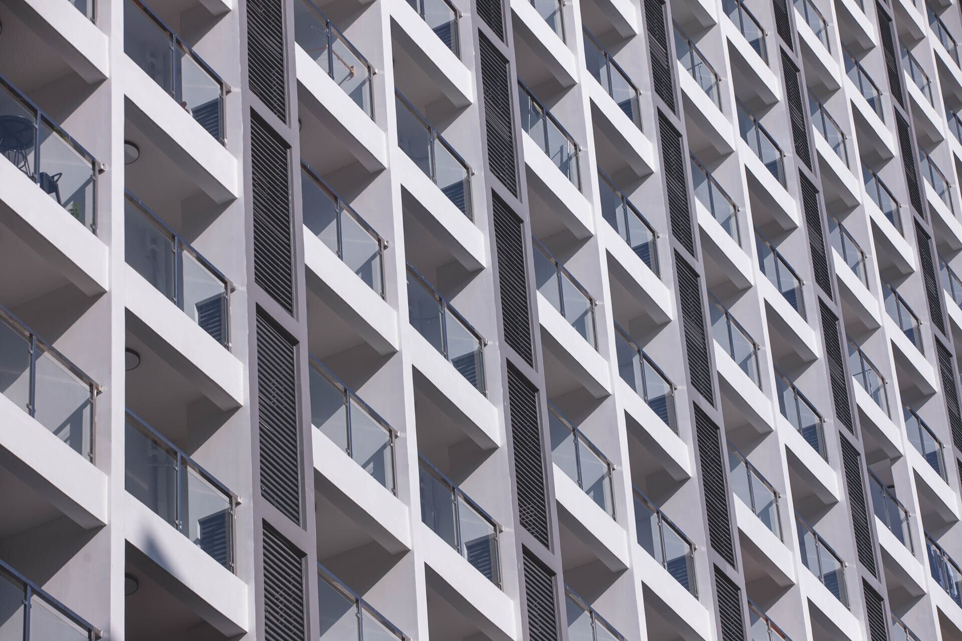 A modern building with a repeating geometric facade, featuring balconies with glass railings and ventilation grilles.