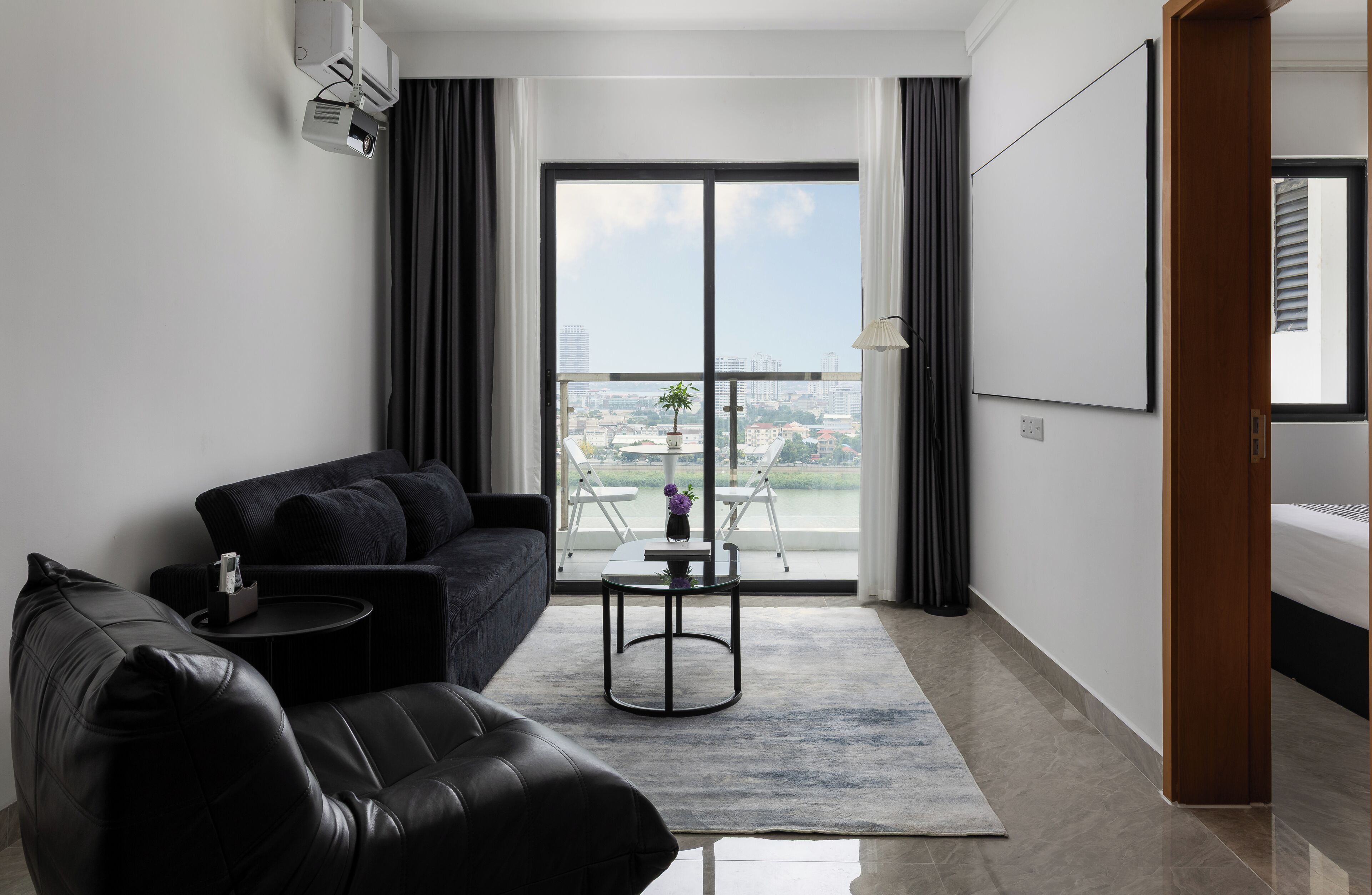 A modern living room with a sofa, a small table, a balcony with a view of the city, and a large window.