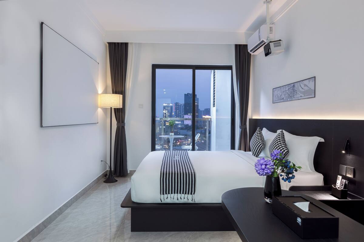 A modern hotel room with a large bed, a bedside table, a lamp, a view of the cityscape, and a mounted projector.