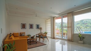 A spacious living room with a sofa, chairs, a coffee table, and a view of a swimming pool through large windows. - Alaya Stays Udgam Vilas 3-BHK With Lift (Dehradun)