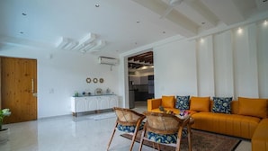 A modern living room with a sofa, chairs, a coffee table, and a kitchen in the background. - Alaya Stays Udgam Vilas 3-BHK With Lift (Dehradun)