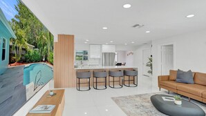 A modern kitchen with a central island, a dining area with bar stools, a pool, and a living room with a sofa and coffee table.