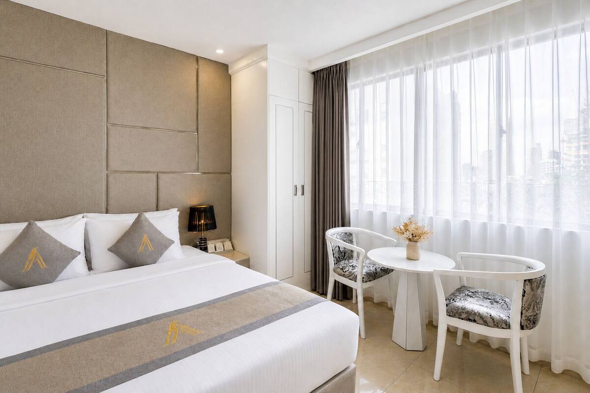 A hotel room with a bed, a nightstand, a lamp, a chair, a small table, and a window with curtains.