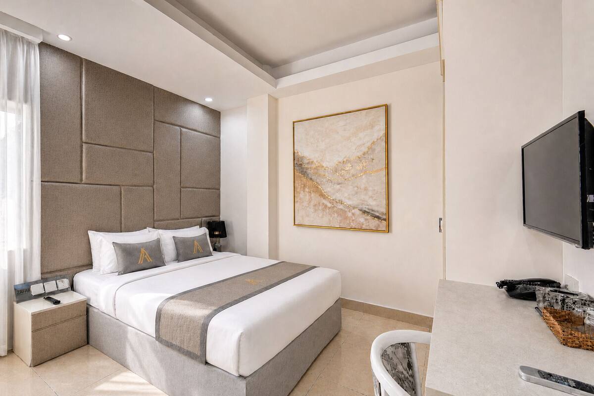 A modern hotel room with a large bed, a flat-screen TV, a desk with a chair, and a large window with sheer curtains.