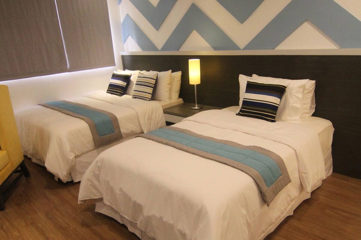 Een hotelkamer met twee bedden, een houten vloer, een wand met een ruiterpatroon en een nachtkastje met een lamp.