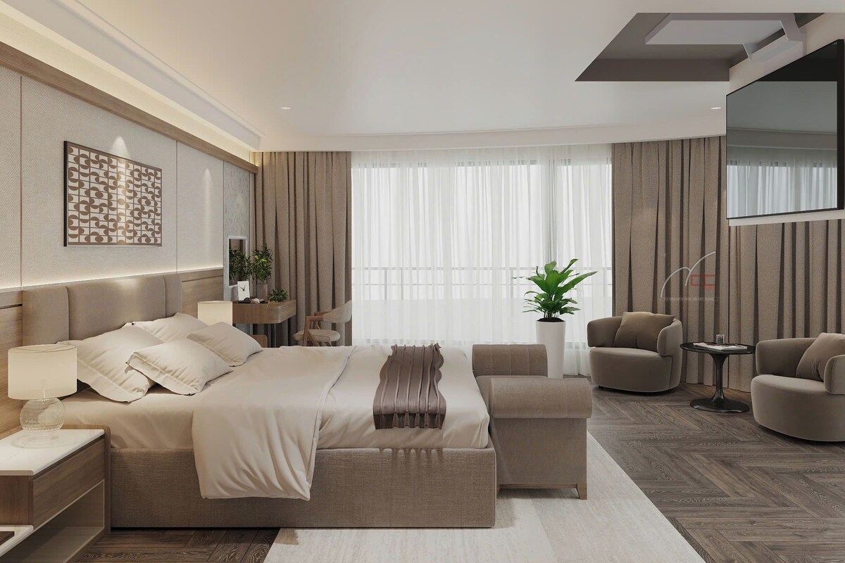 A modern bedroom with a large bed, two armchairs, a side table, a potted plant, and a TV mounted on the wall.