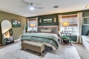 Quarto com cama, um banquinho, mesa de cabeceira com abajur, janela com cortinas e espelho com obras de arte na parede.