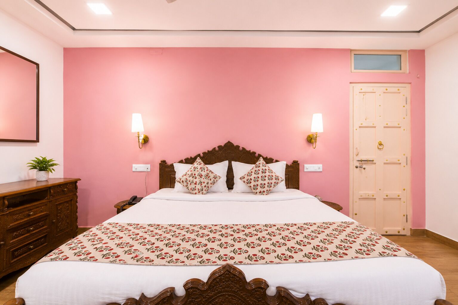 A bedroom with a pink wall, a wooden bed frame, two bedside lamps, a wooden dresser, and a door with a window above it.