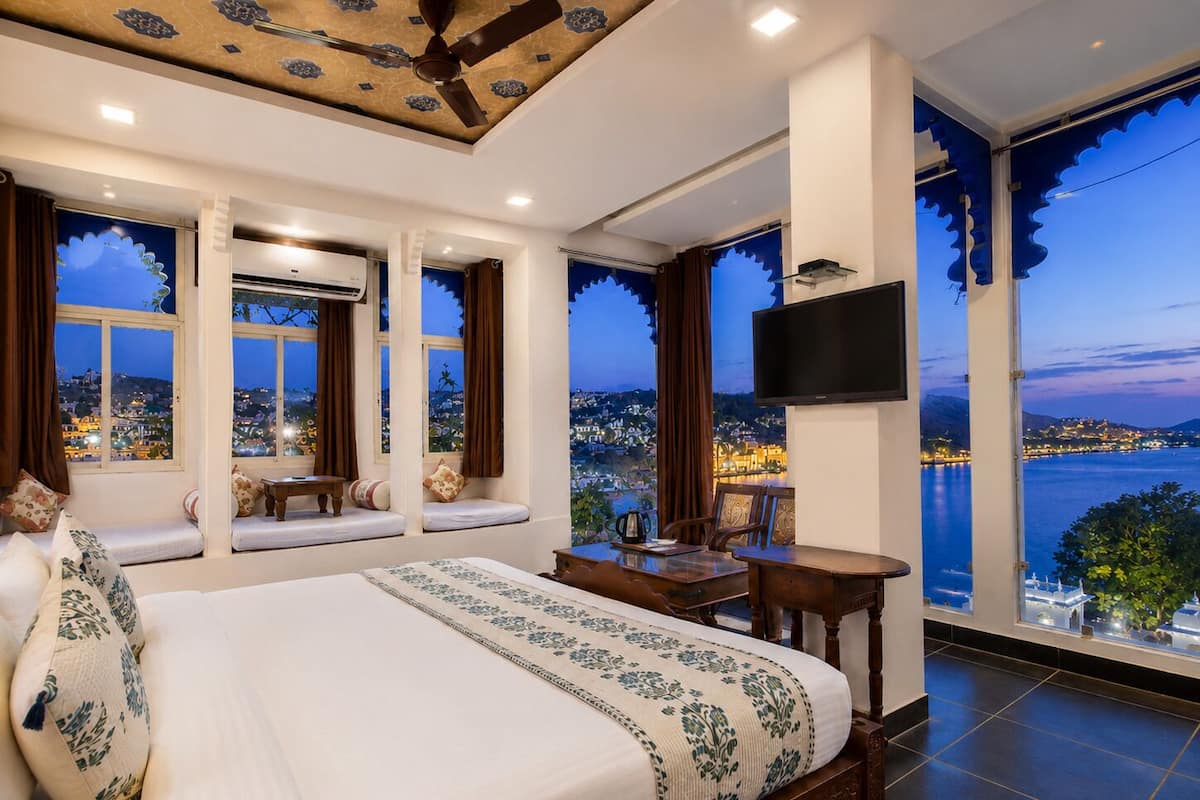 A bedroom with a large bed, a TV, a dining area, and a view of a cityscape at dusk.