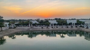 Una masa de agua tranquila que refleja edificios y árboles, rodeada por una cerca blanca y palmeras.