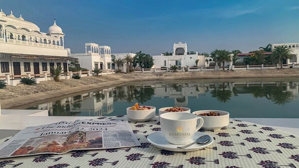 Una mesa con un periódico, tazas y cuencos, con vistas a un cuerpo de agua tranquilo y un edificio blanco con cúpulas.