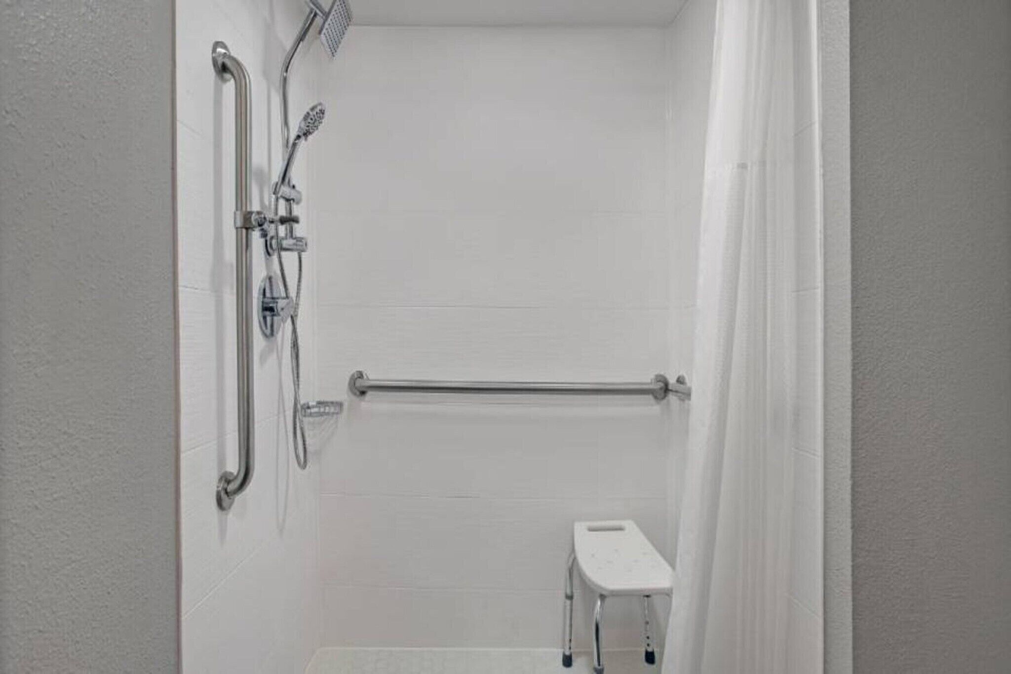 Een witte badkamer met een douche, een greepstang en een klein wit krukje.