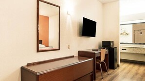 A compact hotel room with a wooden desk, a chair, a flat-screen TV mounted on the wall, and a small kitchenette area.
