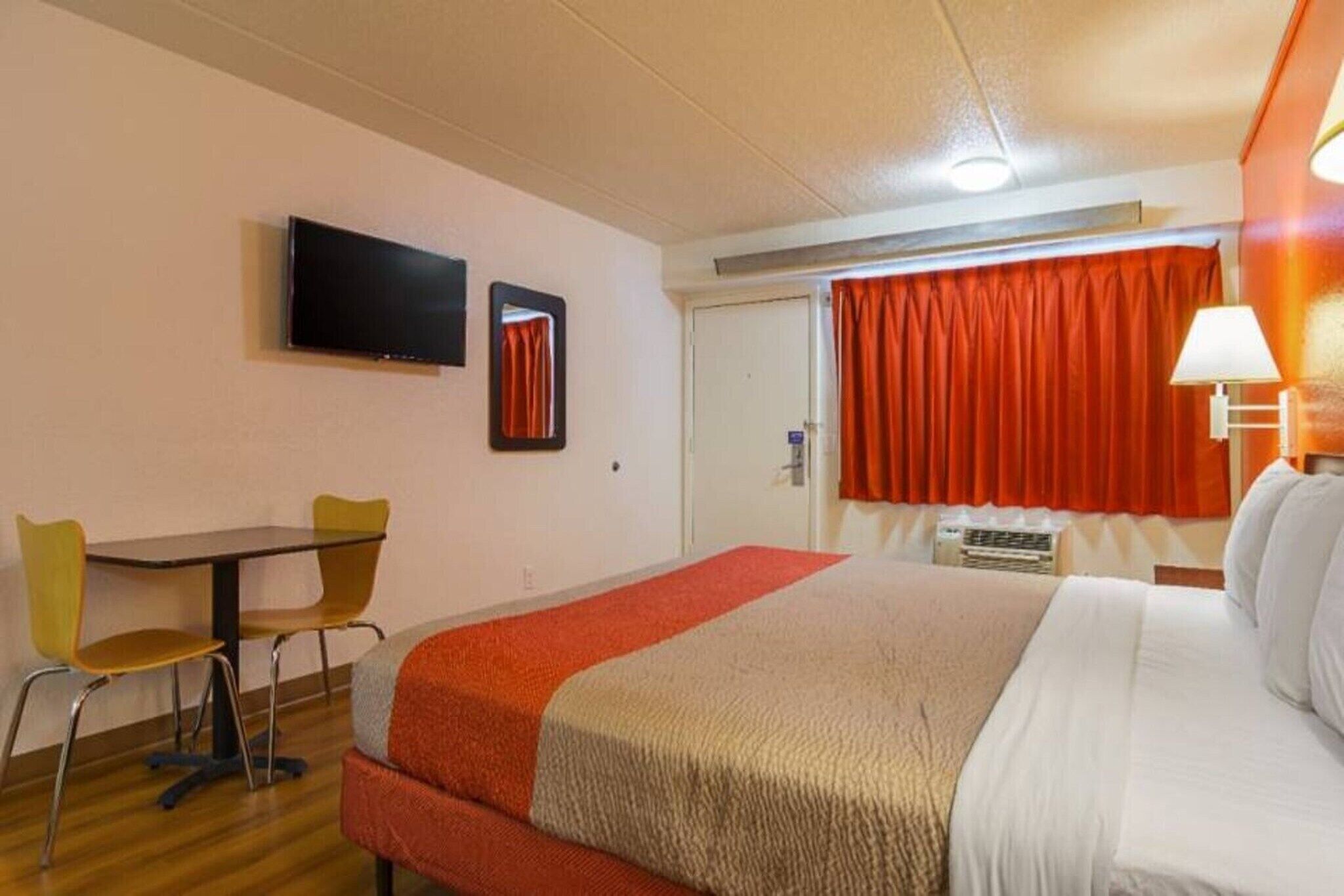 A hotel room with a bed, a desk with two chairs, a television, a window with curtains, and a lamp.