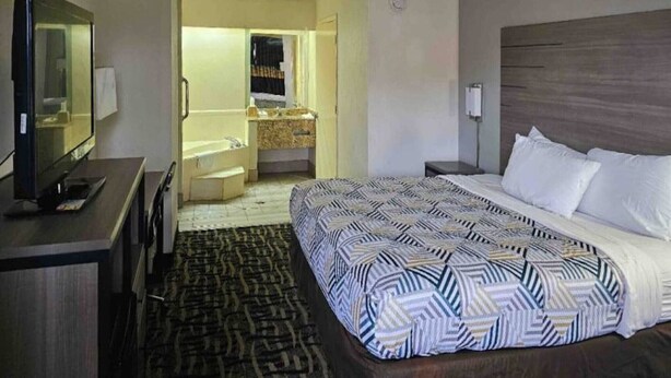 A hotel room with a bed, a TV, a desk, a bathroom with a sink and mirror, and a patterned rug.