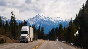 A road with a large truck and a smaller vehicle, surrounded by forest and mountains.
