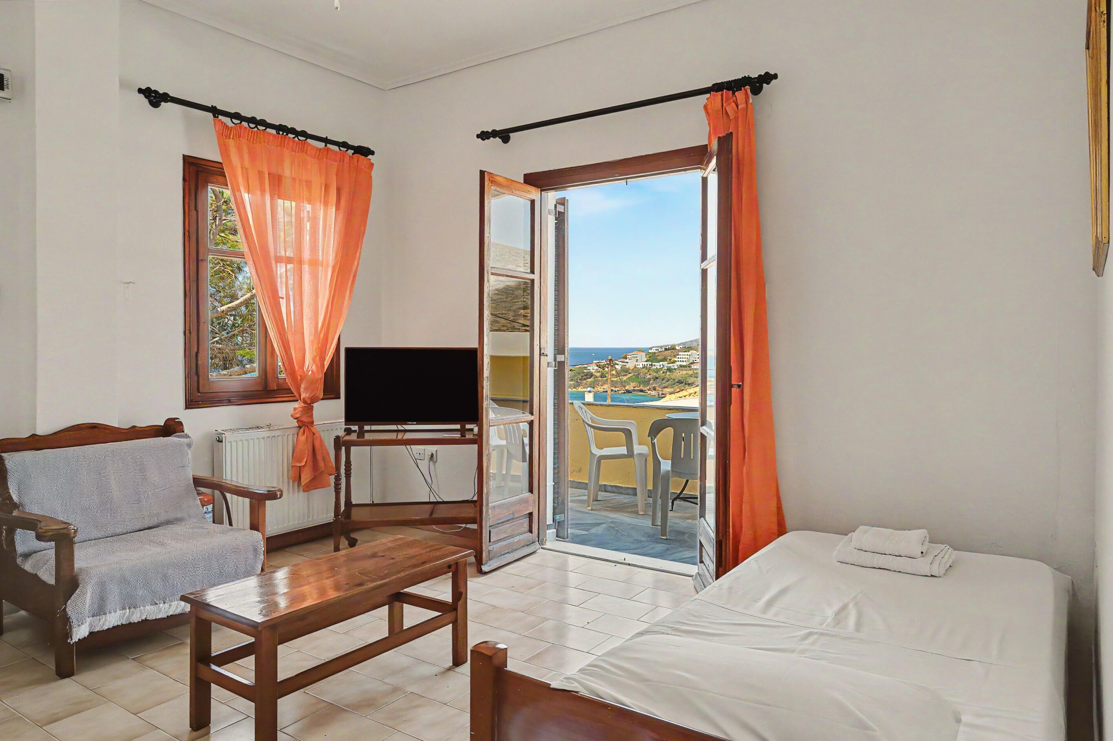 A room with a bed, a TV, a chair, a table, and a view of the sea through an open door.