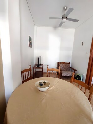 A room with a round table, a ceiling fan, a chair, and a small table with a lamp.