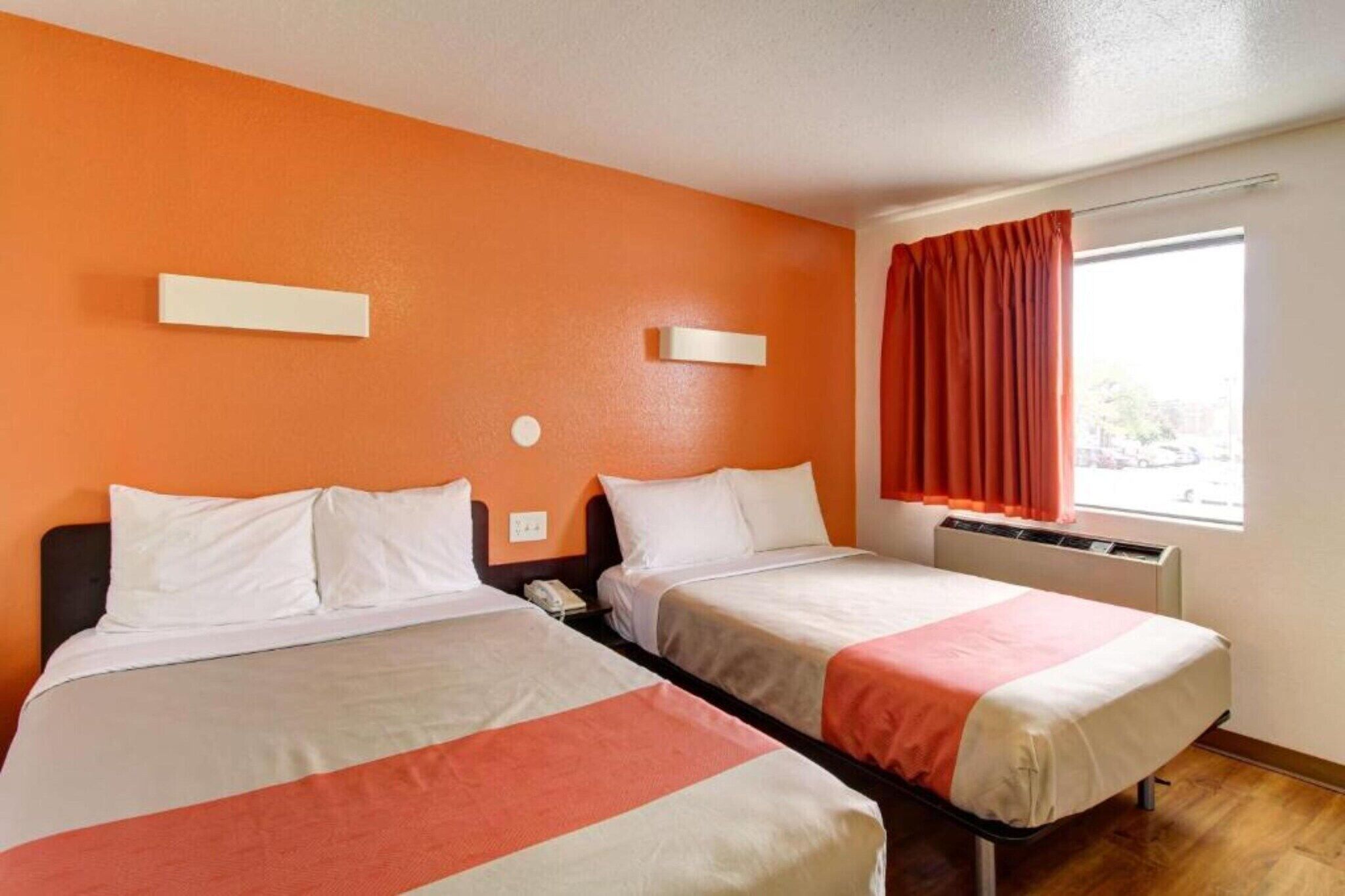 A hotel room with two beds, a window with curtains, and a thermostat on the wall.