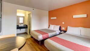 A hotel room with two beds, a small table, a bathroom with a sink, and an orange accent wall. - 2 Full Beds|Smoking,Micfridge (Amherst)
