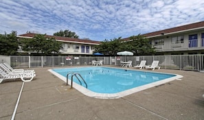 Ein Schwimmbad mit Liegestühlen, ein eingezäunter Bereich und ein Gebäude mit mehreren Balkonen im Hintergrund.