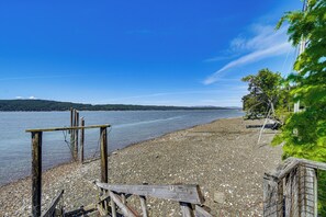 Una spiaggia con sassolini, strutture in legno, cielo sereno e colline in lontananza.