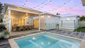 Un patio trasero con piscina, mobiliario de jardín y luces colgantes.