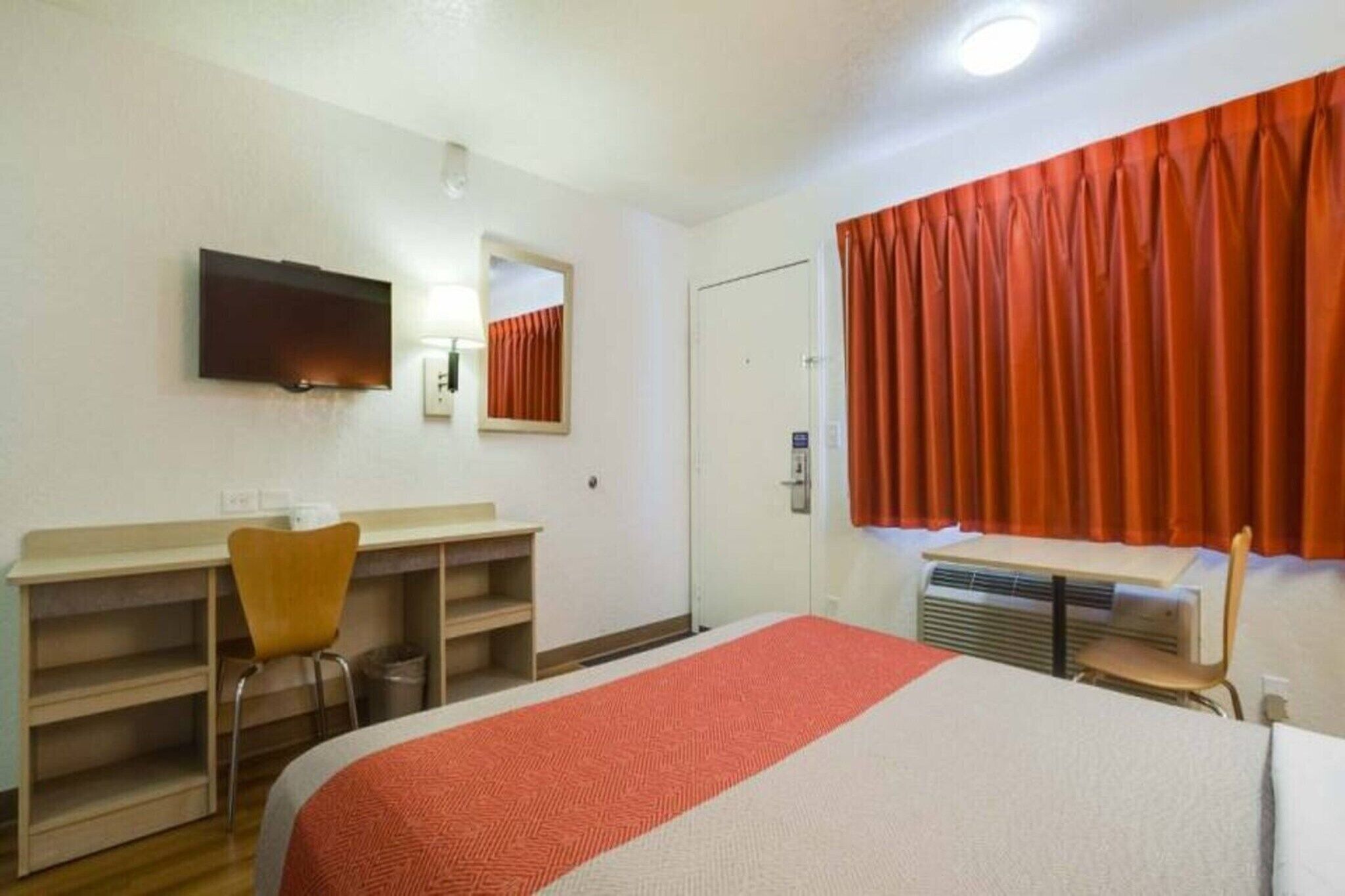 Una camera d'albergo con un letto, una scrivania, una sedia, una televisione, uno specchio, una porta e una finestra con tende.