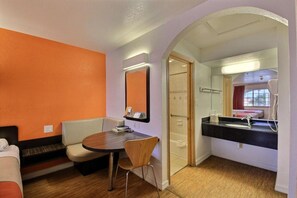 A hotel room with a dining table, a bench, a shower area, and a bathroom with a sink and mirror.