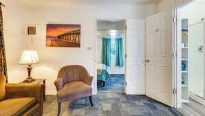 Hotellihuone, jossa on tuoli, lamppu, taulu ja avoinna oleva ovi, joka johtaa toiseen huoneeseen.