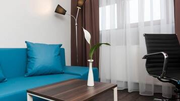 Ein modernes Zimmer mit einem blauen Sofa, einer weißen Vase mit Pflanze, einem dunklen Holztisch, einem schwarzen Bürostuhl und einer Stehlampe.