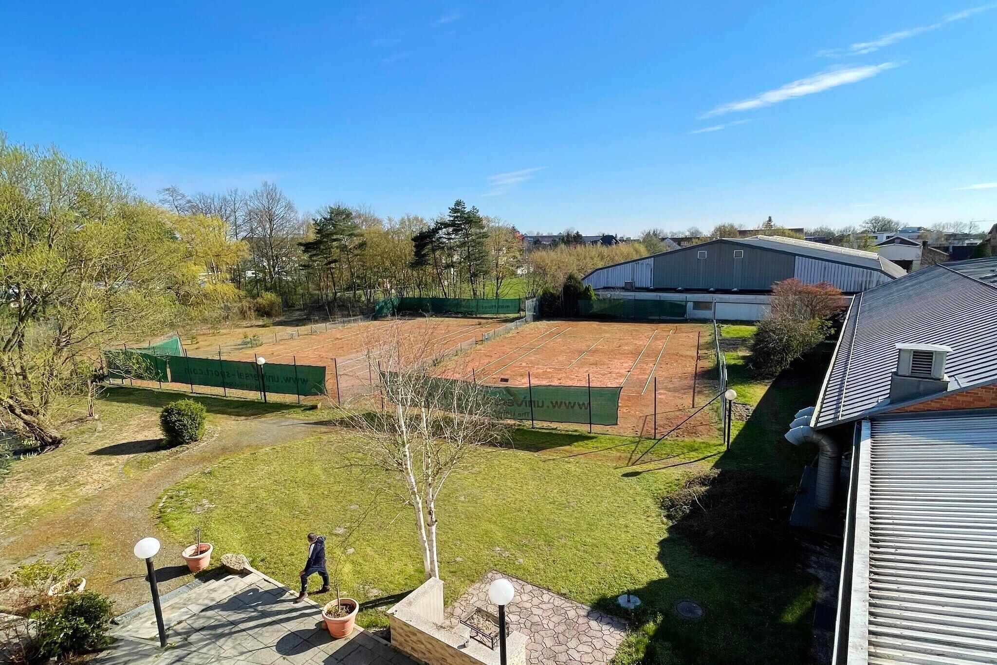 A tennis court surrounded by a fenced area, a building with a sloped roof, and a clear sky.