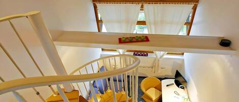 Una escalera con barandilla blanca, una pequeña mesa con un jarrón y un girasol, y una estantería con manzanas.