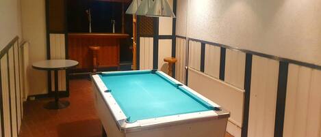 A pool table in a room with a bar area, a stool, and a hanging light fixture.