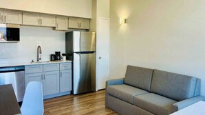 A compact kitchen with a refrigerator, microwave, and coffee maker. There's a gray sofa and a white chair in the living area.