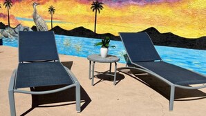 Two lounge chairs, a small table with a potted plant, and a mural of a sunset over water.