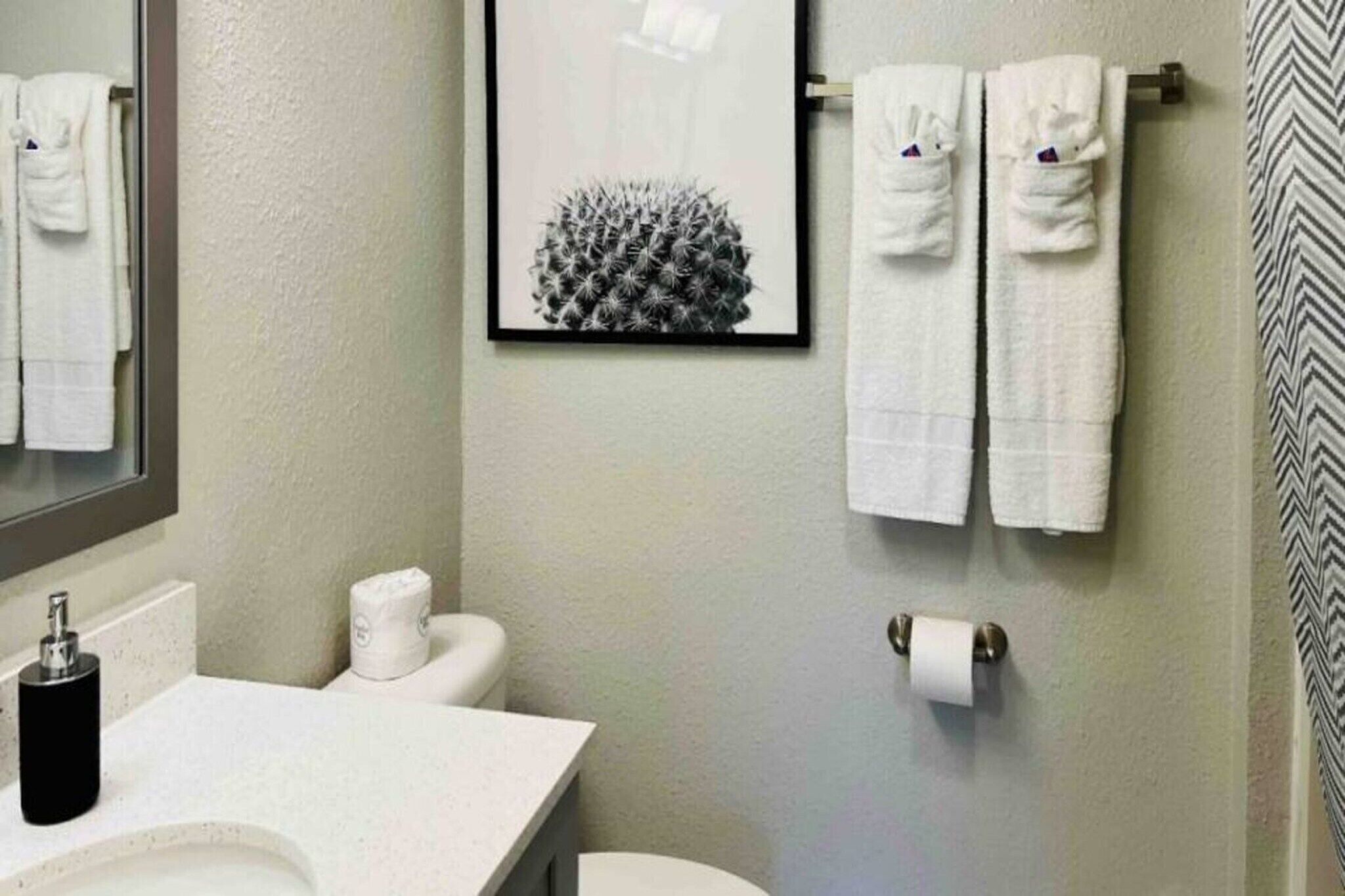 Un baño con inodoro, lavabo, un cuadro con motivos de cactus y dos toallas en un escurreplatos.