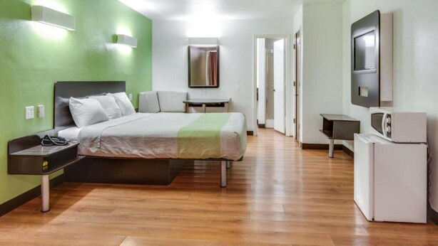 A hotel room with a bed, bedside table, TV, and a small kitchenette. - 1 King Bed | Smoking, Kitchen, Sofa-Bed (Longview)