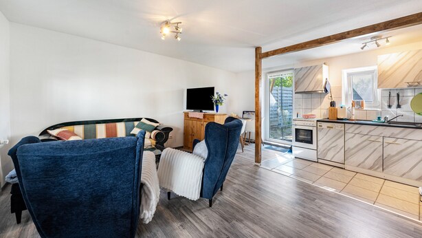 A modern living room with a sofa, a television, and a wooden dining table. A kitchen with a stove, sink, and cabinets is visible through an open doorway. - Newly Built Holiday Apartment with Terrace (Klein Wittensee)
