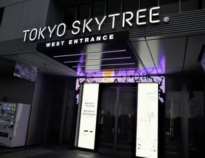 Der Westeingang des Tokyo SkyTree mit modernem Design, darunter beleuchtete Schilder und ein Getränkeautomat.