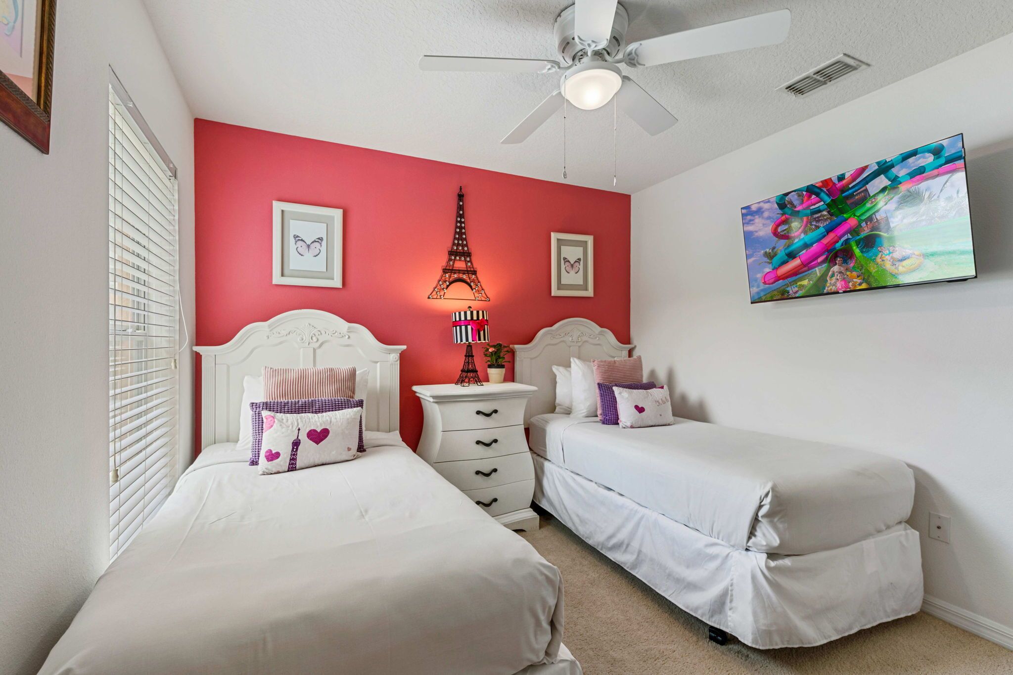 Une chambre avec deux lits, un ventilateur de plafond, un téléviseur fixé au mur et un mur d’accent de couleur rouge.